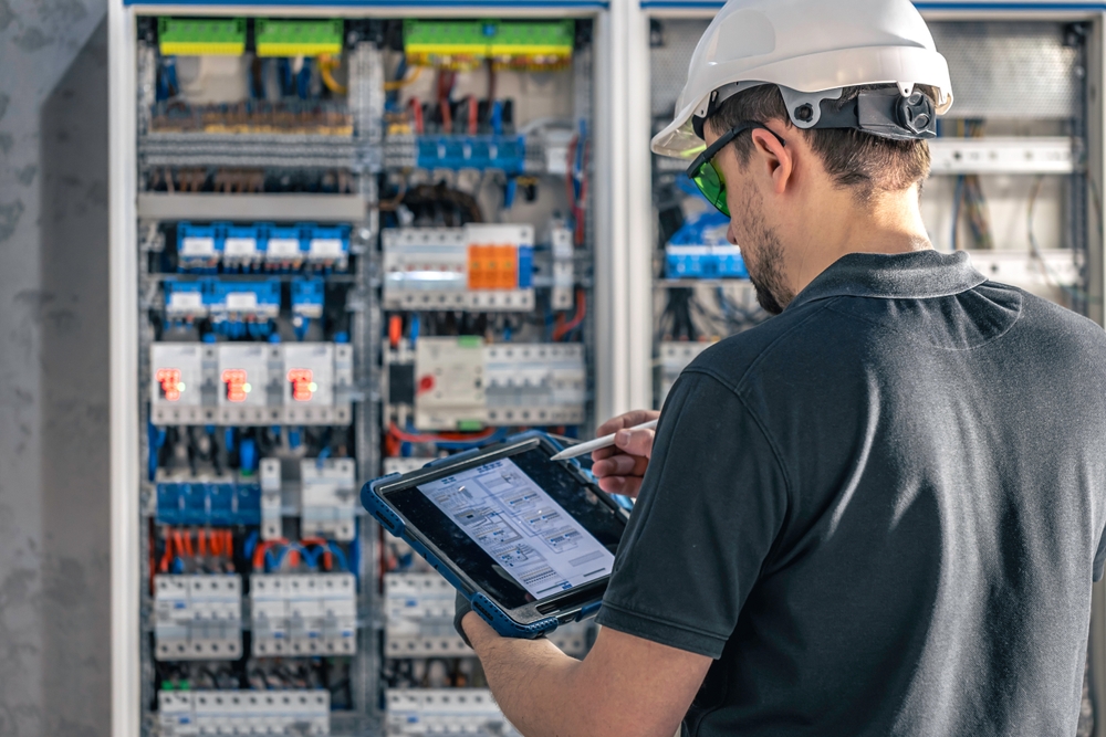 Photo of an electrician with a helmet and goggles examining a switchboard with fuses, while using a tablet.